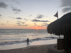 Escena de playa con atardecer, olas y una estructura de techo de paja.