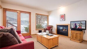 A living room with a red sofa, wooden coffee table, fireplace, television, and large windows with blinds. - Storm Watch Condominiums - Sw205 (Steamboat Springs)