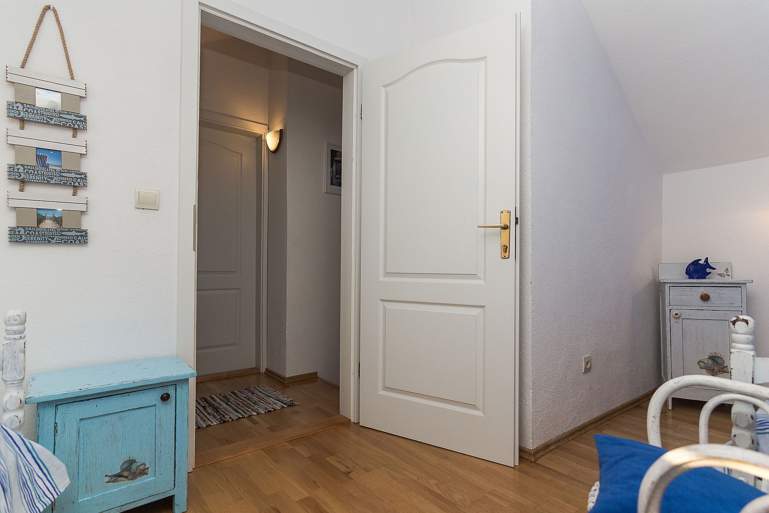 A room with a wooden floor, a white door, a small blue cabinet, and a white chair with a blue cushion.
