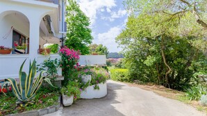 A white building with a covered porch, potted plants, and a pathway leading to a road. - Apartment Marija (Palit)