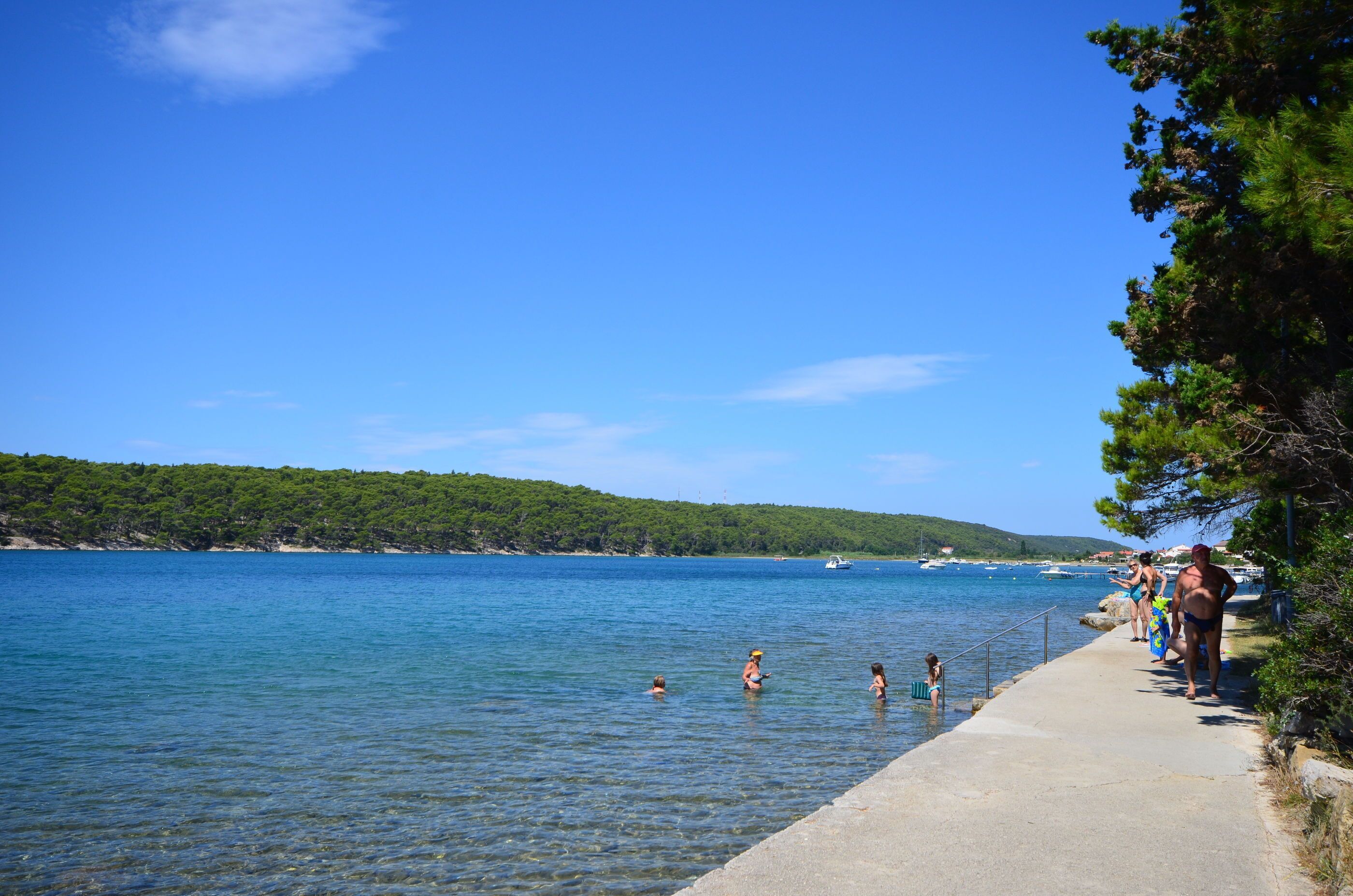 A coastal walkway with clear blue water, a concrete path, and a forested hillside in the background.