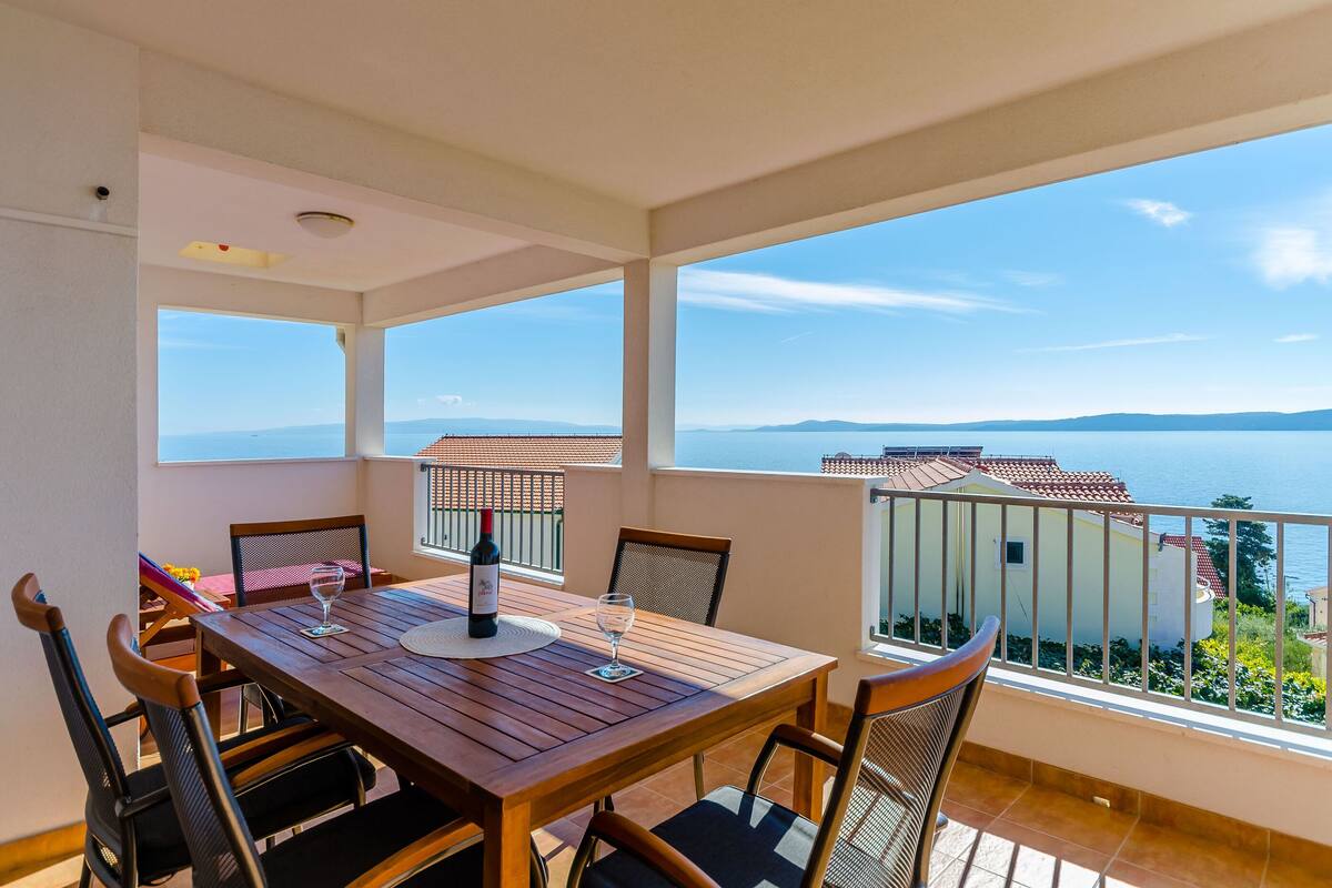 A balcony with a wooden table and chairs, a bottle of wine, and a glass. The balcony overlooks a scenic view of the sea and distant mountains.