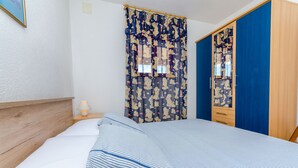 A neatly made bed with striped bedding, a wooden headboard, a bedside lamp, a patterned curtain, and a blue wardrobe with a mirror. - Apartments Jurko (Okrug Gornji)