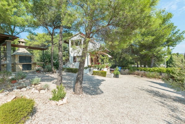 Une maison avec une cheminée en pierre, entourée d’une allée en gravier et de diverses plantes.