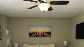 Une chambre à coucher comprenant un lit, deux tables de chevet, un ventilateur de plafond, un tableau et un téléviseur fixé au mur.