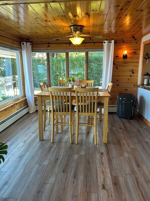 A wooden dining area with a table and chairs, large windows with curtains, and a ceiling fan.