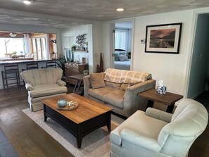 A living room with a wooden coffee table, two sofas, a small side table, and a dining area with chairs and a kitchen counter.