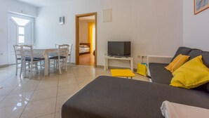 Ein modernes Wohnzimmer mit einer Couch, einem Fernseher, einem Esstisch mit Stühlen und einem kleinen, gelben Hocker.