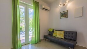 Ein Zimmer mit grauem Sofa, grünen Vorhängen, Fliesenboden und einem Bild an der Wand.