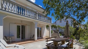 Ein überdachter Außenbereich mit Tischen und Stühlen, eine Treppe zu einem zweiten Stockwerk und ein Garten mit Pflanzen und einer Pergola.