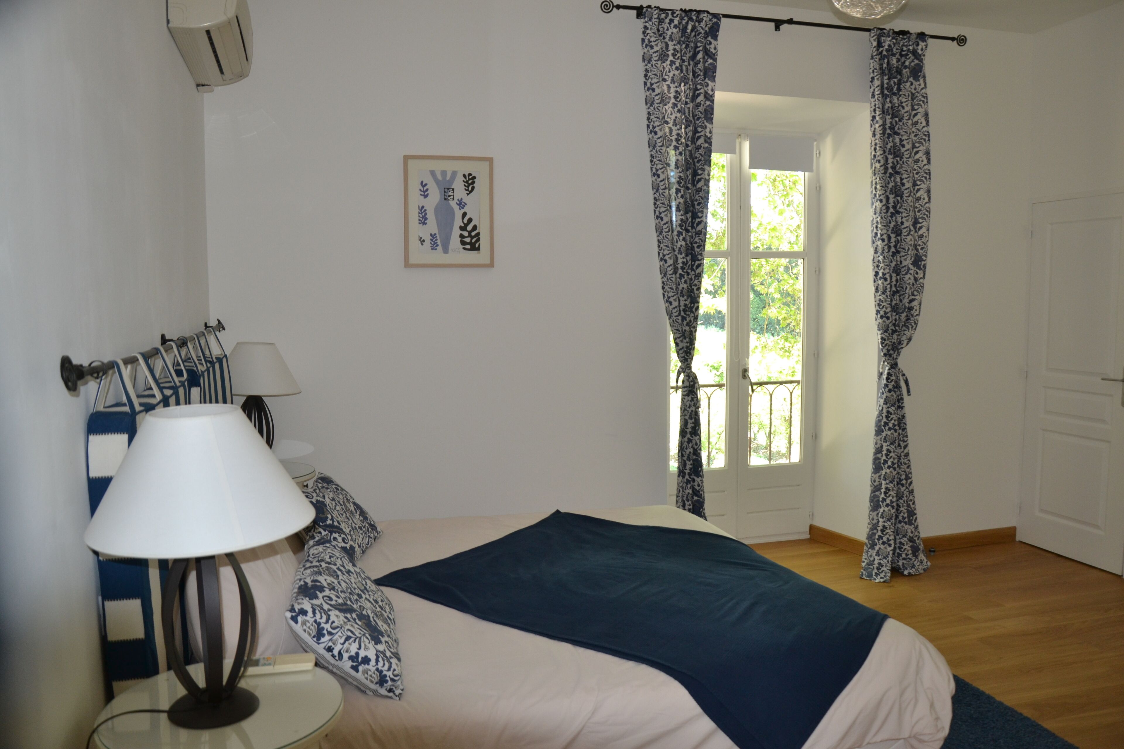 Un dormitorio con cama, mesita de noche con lámpara, cortinas estampadas y ventana con vista a follaje.
