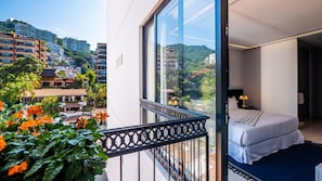 Un balcone con vista sulla città, un letto con materasso bianco e un cuscino azzurro, e un tavolino con una lampada.