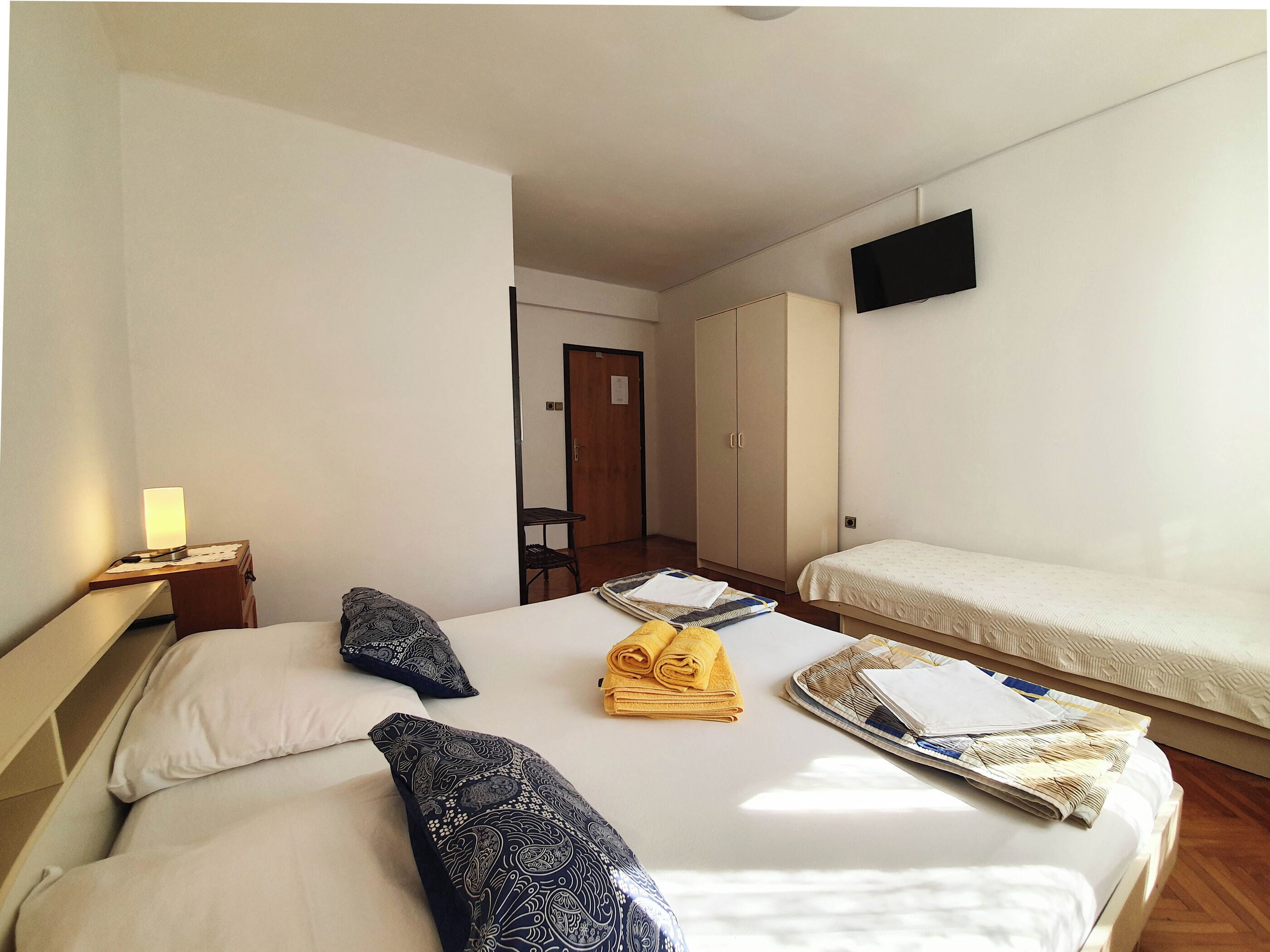 Quarto de hotel com duas camas, uma televisão fixada na parede, uma escrivaninha com abajur e um guarda-roupa.
