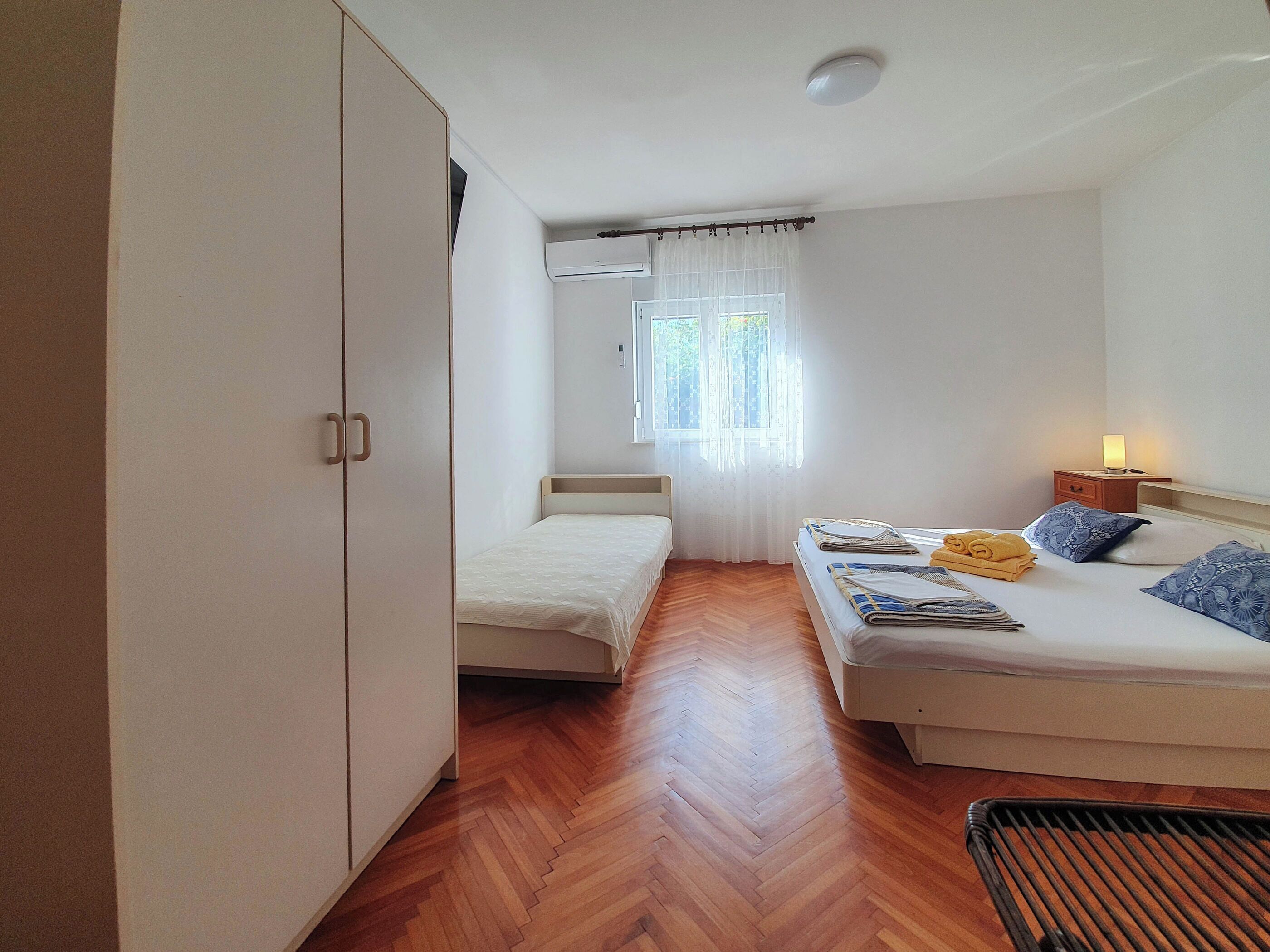 A small, well-lit room with two beds, a wardrobe, and a window with sheer curtains.
