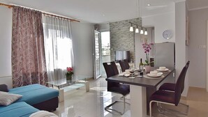 A modern living room with a blue sofa, a dining area with a table set for two, and a kitchen visible through an open door. - Apartments Ruzica (Banjol)