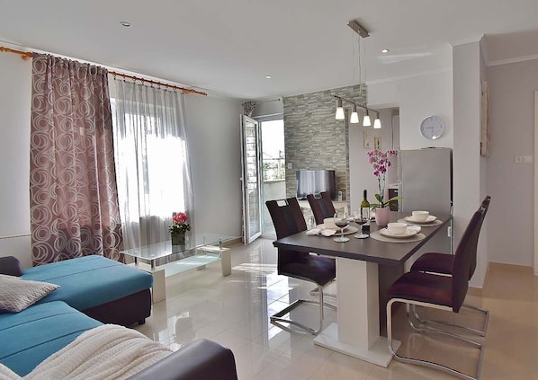 A modern living room with a blue sofa, a dining area with a table set for two, and a kitchen visible through an open door. - Apartments Ruzica (Banjol)