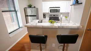 Una cocina con armarios blancos, un refrigerador, un microondas, un horno y dos taburetes negros.