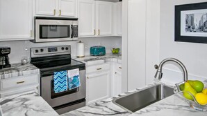 Una cocina moderna con armarios blancos, un horno de acero inoxidable, un microondas y un lavabo con un bol de fruta.