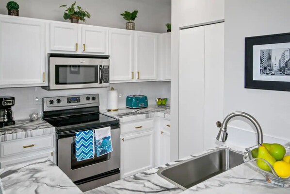 A modern kitchen with white cabinets, a stainless steel oven, a microwave, and a sink with a bowl of fruit.