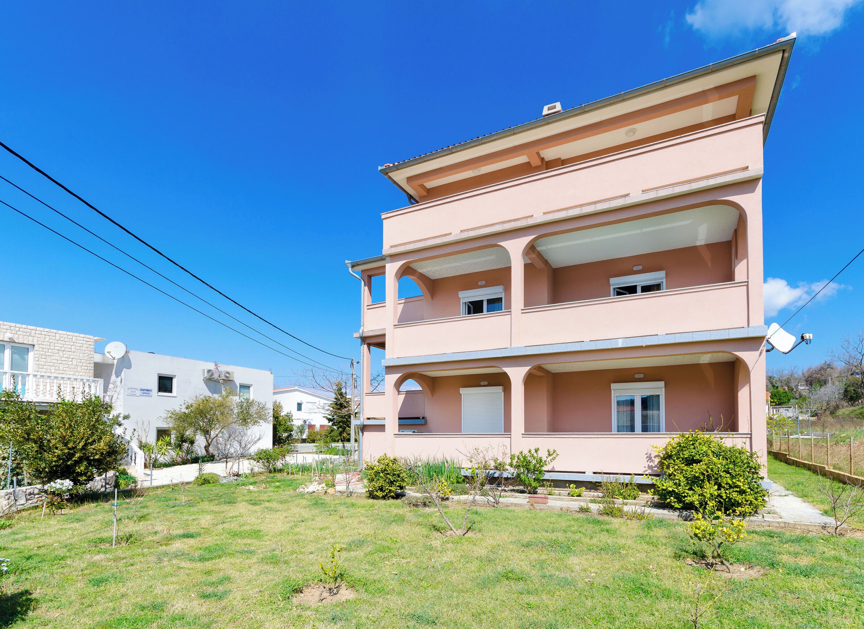 A multi-story residential building with a flat roof, balconies, and a garden area in front.
