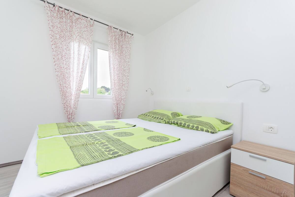 Un dormitorio con cama, mesita de noche, ventana con cortinas y vistas al exterior.