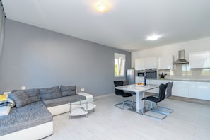 A modern living space with a grey sofa, a glass dining table, and a kitchen area with white cabinets and stainless steel appliances.