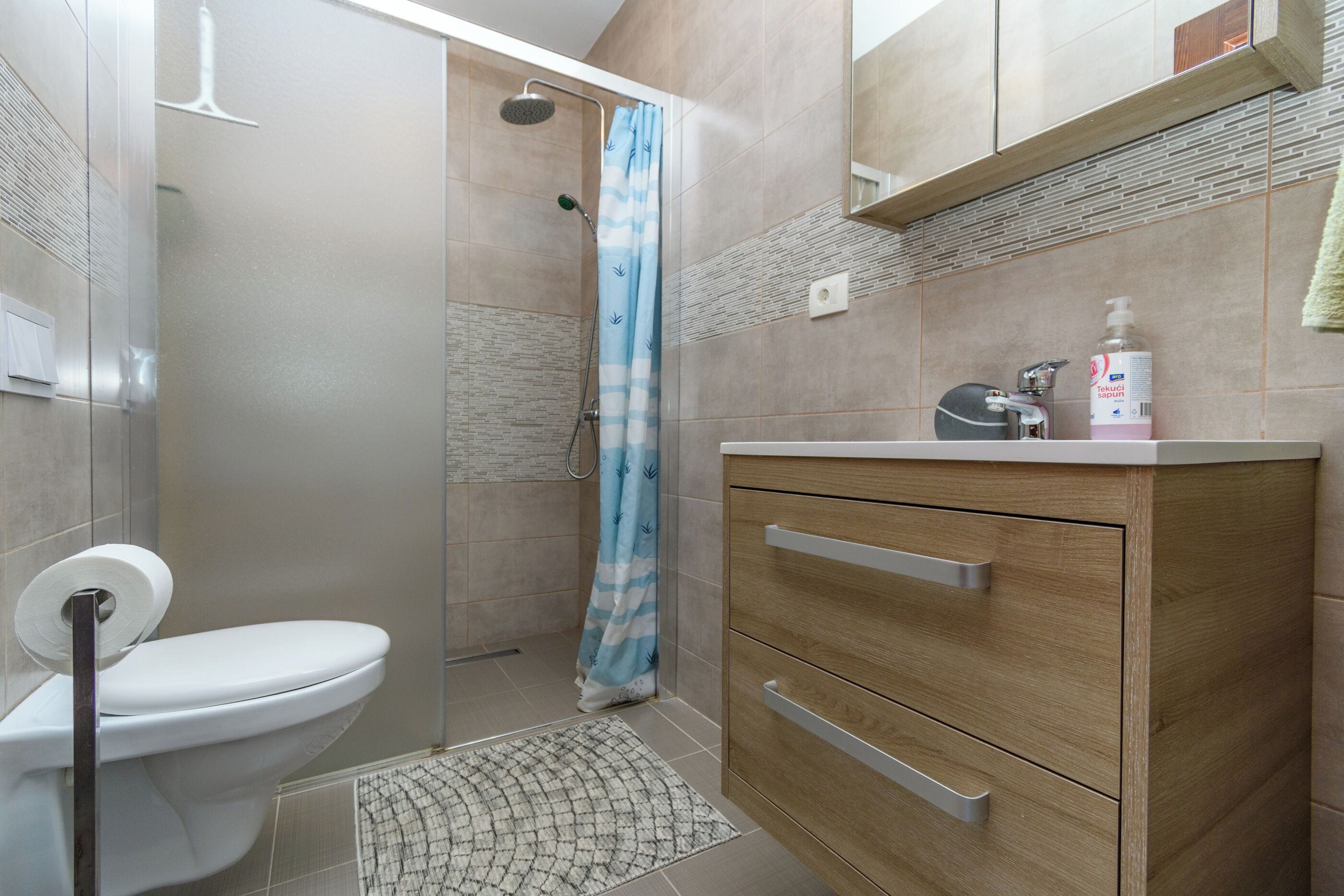 A bathroom with a toilet, a shower with a curtain, a sink with a soap dispenser, and a wooden cabinet.