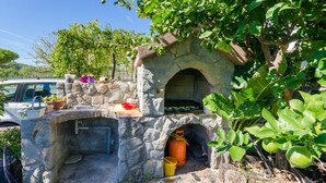 A stone-built outdoor oven with a brick roof, surrounded by greenery and a parked car. - Apartments Elvira (Lopar)