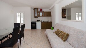 A compact living space with a kitchenette, a dining area with a glass table and chairs, and a sofa with a patterned cushion. - Apartments Elvira (Lopar)