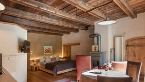 Une chambre à coucher au style rustique, avec des poutres apparentes, un lit doté d’une tête de lit sombre, une chaise rose, une petite table sur laquelle est posé un vase de fleurs et une cheminée.