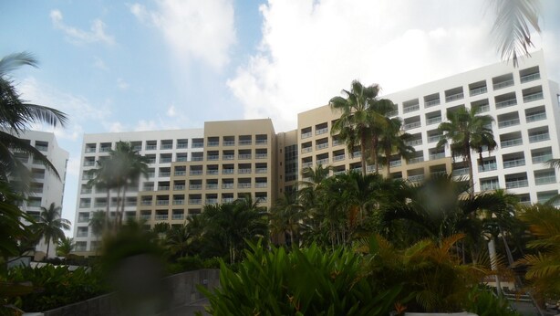 Un hotel de varios pisos, un jardín ajardinado con palmeras y un cielo despejado.