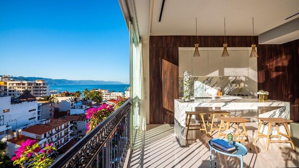 Un balcone con vista sul mare e sugli edifici, un angolo cottura con un piano di lavoro in marmo e sgabelli di legno.