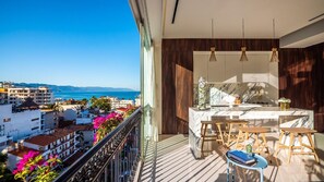 A balcony with a view of the sea and buildings, a kitchen area with a marble countertop, and wooden stools.
