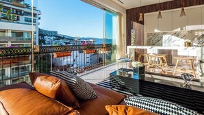 Una sala de estar moderna con un ventanal que ofrece vistas a la ciudad, un balcón con panorama y una zona de cocina.
