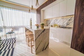 A modern kitchen with marble countertops, a central island with bar stools, and a view of the outdoors through large windows. - PIER 57 - 508 | Modern 2 BR with Great View in PV (Puerto Vallarta)