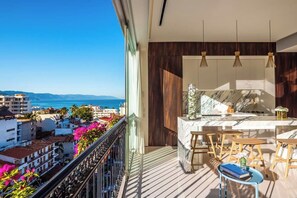 A balcony with a view of the sea and buildings, a kitchen with wooden cabinets and pendant lights, and a small table with books and a vase. - PIER 57 - 508 | Modern 2 BR with Great View in PV (Puerto Vallarta)
