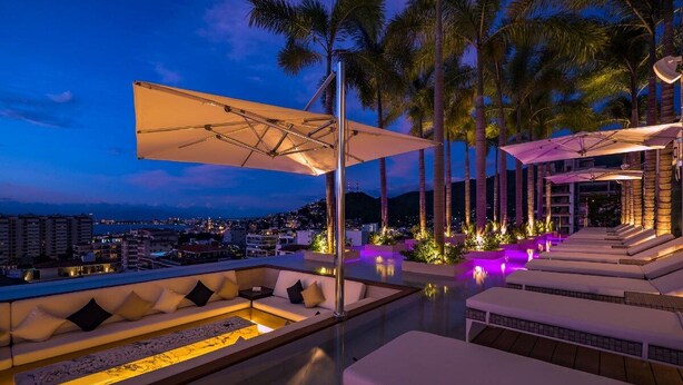Zona de piscina en la azotea con sillones reclinables y vista a la ciudad al anochecer.