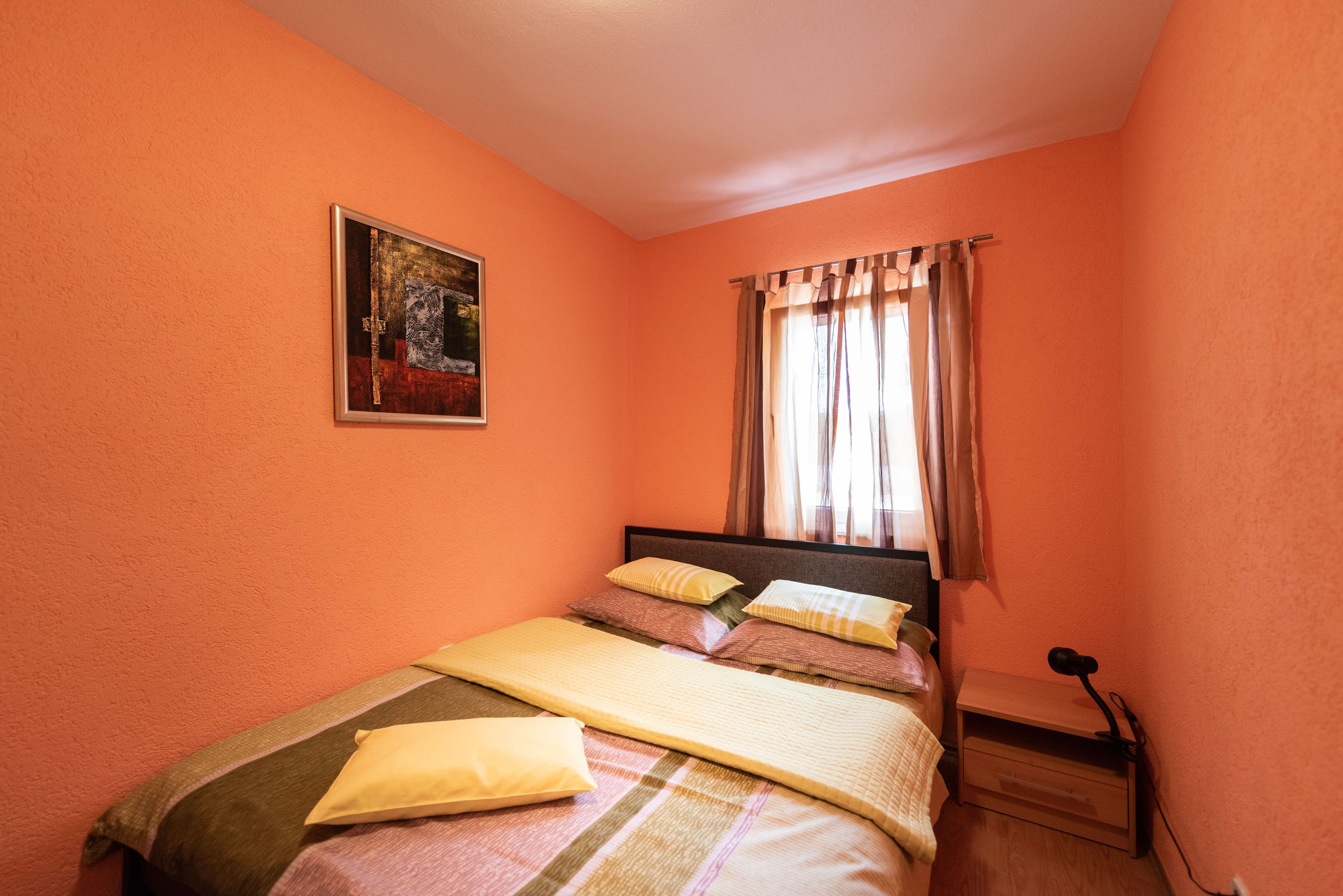 Une chambre à coucher avec un lit, une table de chevet, une lampe, une fenêtre avec des rideaux et un tableau au mur.