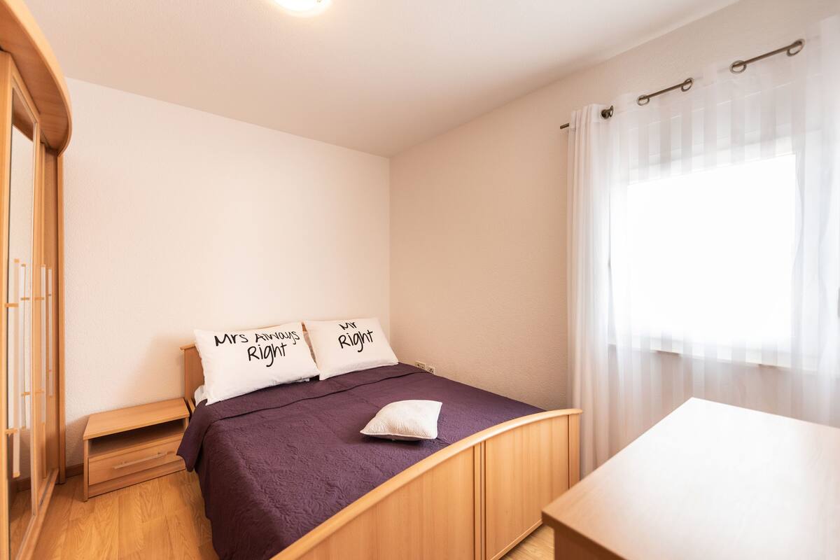A bedroom with a bed, a wooden nightstand, a small table, and a window with sheer curtains.