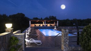 Area piscina illuminata di notte, con solarium in pietra, lettini e una zona relax coperta sotto un cielo stellato.