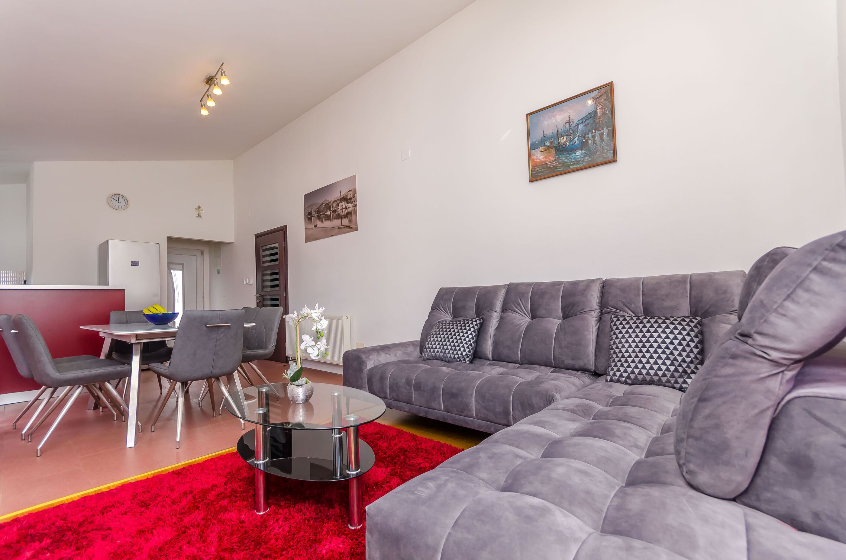 Una sala de estar moderna con un sofá gris, una mesa de centro de vidrio y una alfombra roja. Hay un comedor con una mesa y sillas, y una cocina con un refrigerador y un microondas.