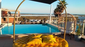 A poolside area with a yellow and white striped lounge chair, a glass-enclosed cabana, and a view of the beach and ocean. - Stylish apartment with amazing views of the Ria Formosa & the sea in Fuseta. (Fuseta, Moncarapacho.)