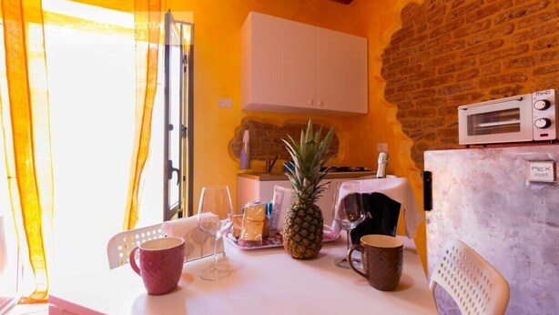A kitchen with a brick wall, white cabinets, a microwave, and a table set with a pineapple, glasses, and a mug. - Patty Studio 'Casa a Chiazzetta' (Castelbuono)