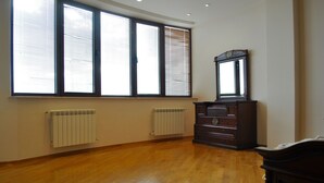 A room with wooden flooring, a large window with blinds, a radiator, and a dark wooden dresser with a mirror. - Apartment on Ismayil bay Qurtqashinli 50 (Baku)