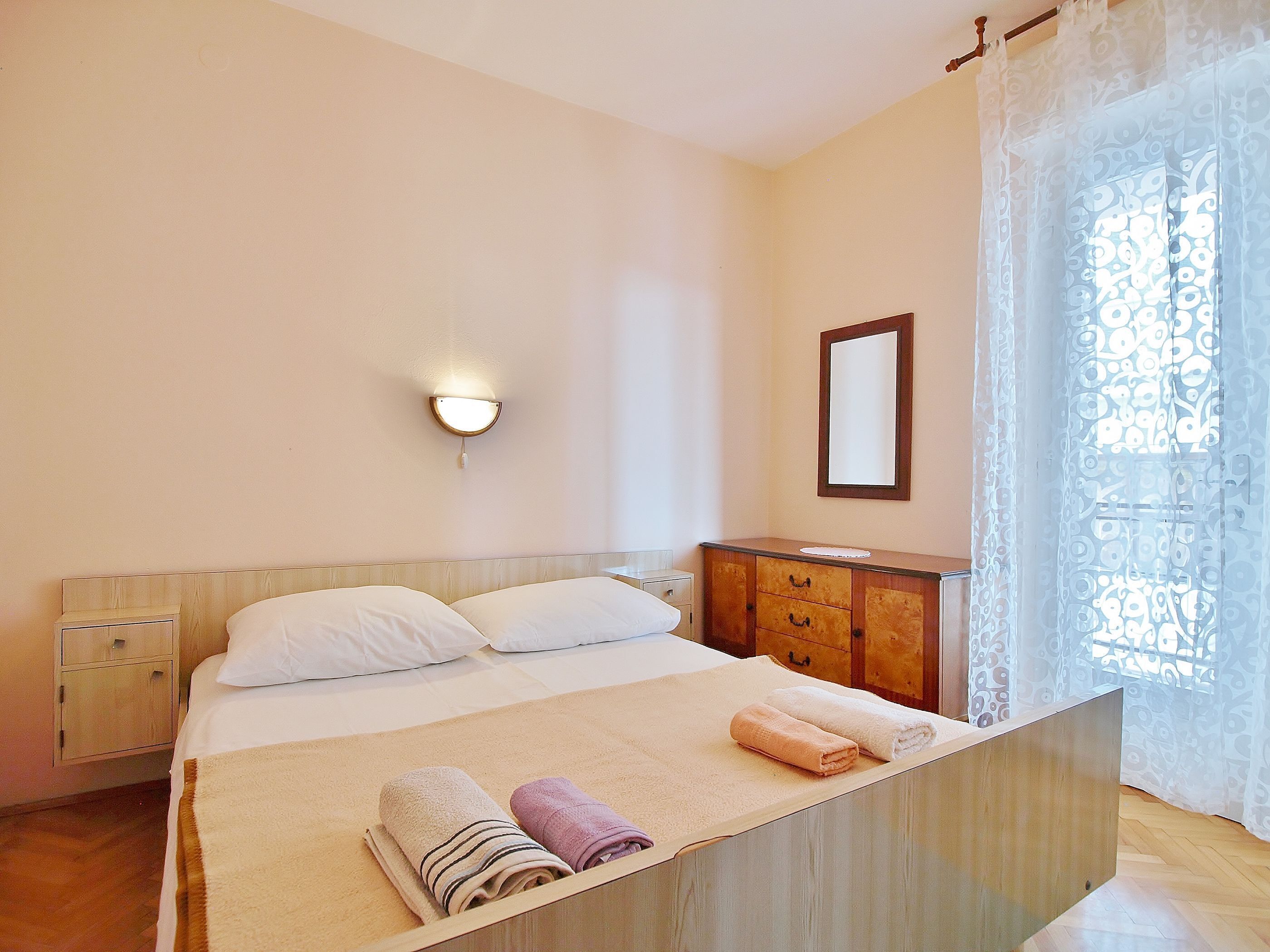 Een hotelkamer met een bed, nachtkastje, houten dressoir, spiegel en raam met dunne gordijnen.