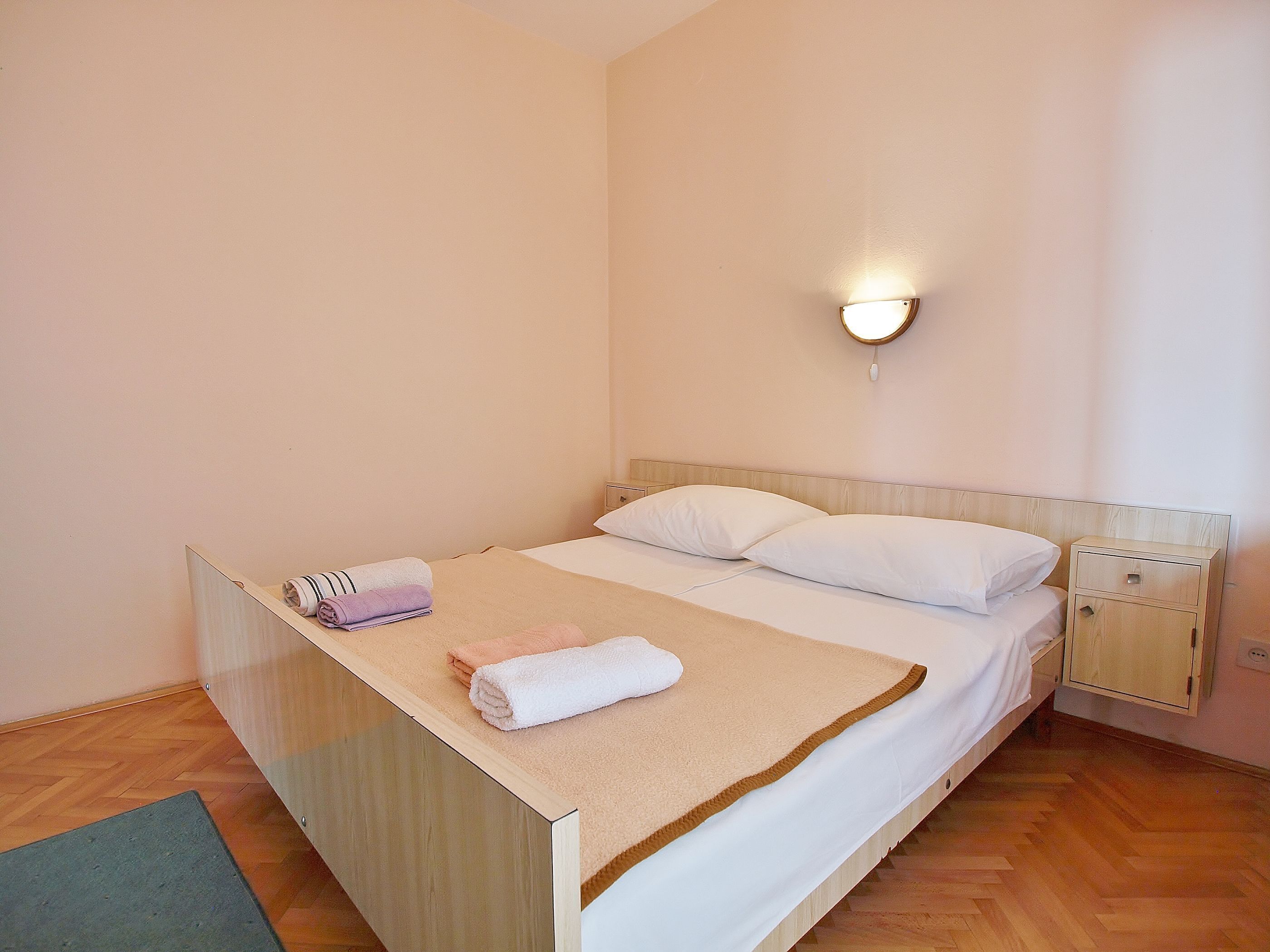 A neatly made bed with a beige blanket and white pillows, a wooden headboard, a bedside table with a lamp, and a small wooden cabinet.