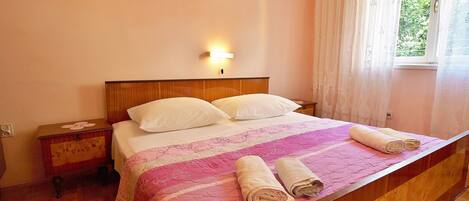 A neatly made bed with a pink and white quilt, two pillows, and rolled towels on a wooden bed frame in a room with a wooden nightstand and a wall-mounted light.