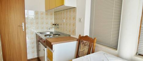 A compact kitchen with a sink, stove, and wooden cabinets. There is a dining table with a blue ashtray and a chair with a floral pattern.