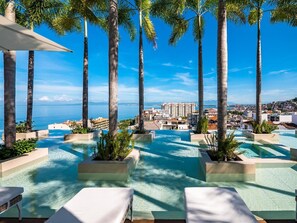 A rooftop infinity pool with palm trees, lounge chairs, and a view of the city and sea.