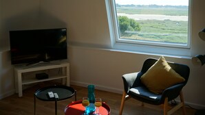 A living room with a television on a white stand, a chair with a yellow pillow, a small table with a remote control, and a window with a view of a field and water.